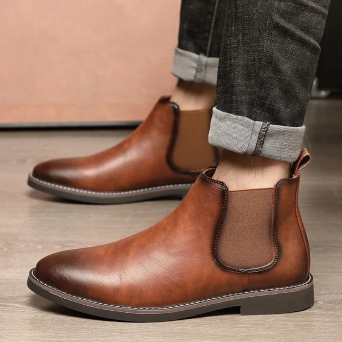 Men's black leather Chelsea boots on a wooden floor, lighting emphasizes texture.
